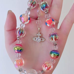 May include: A colorful beaded keychain with a clear, iridescent charm and a small, gold charm with a clear crystal in the center. The keychain is made of clear, iridescent beads, and some of the beads have a rainbow effect.