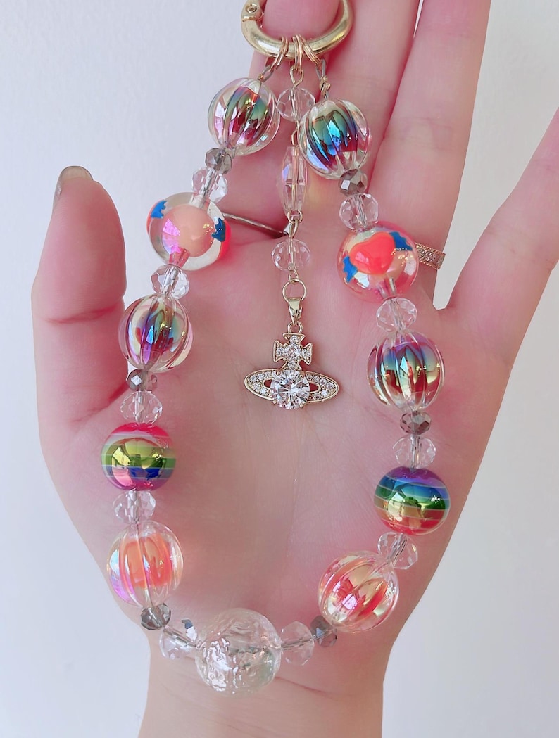 May include: A colorful beaded keychain with a clear, iridescent charm and a small, gold charm with a clear crystal in the center. The keychain is made of clear, iridescent beads, and some of the beads have a rainbow effect.