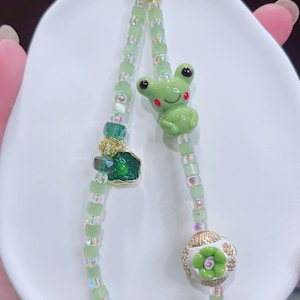 May include: A green beaded keychain featuring a frog charm, a green gemstone, and a white and gold floral bead. The frog charm has red cheeks and black eyes. The keychain is made of small, square green beads and iridescent beads.