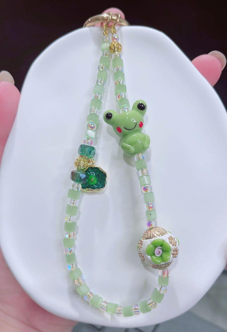 May include: A green beaded keychain featuring a frog charm, a green gemstone, and a white and gold floral bead. The frog charm has red cheeks and black eyes. The keychain is made of small, square green beads and iridescent beads.