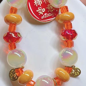 May include: A beaded bracelet with iridescent orbs, orange and yellow beads, and red faceted beads. A red tag with gold lettering and a small orange charm are attached. The bracelet is designed with a mix of shapes and colours.