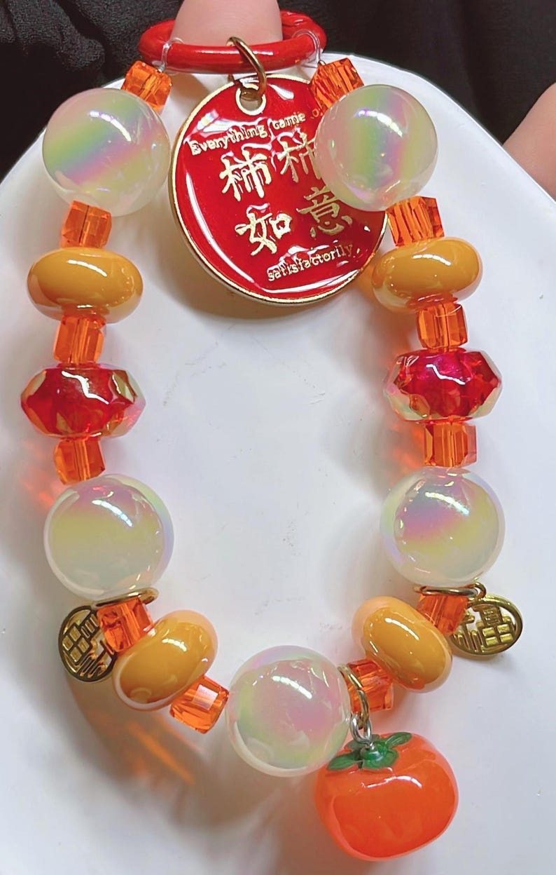May include: A beaded bracelet with iridescent orbs, orange and yellow beads, and red faceted beads. A red tag with gold lettering and a small orange charm are attached. The bracelet is designed with a mix of shapes and colours.