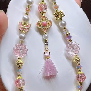 May include: An elaborate necklace with pearls, pink beads, and gold detailing. The design incorporates a butterfly, flower, and tassel pendant. The necklace is presented on a white surface.