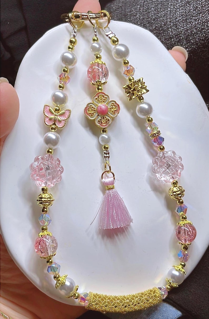 May include: An elaborate necklace with pearls, pink beads, and gold detailing. The design incorporates a butterfly, flower, and tassel pendant. The necklace is presented on a white surface.