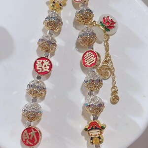 May include: A gold bracelet with decorative beads and charms. The bracelet features round, textured beads interspersed with smaller, crystal-studded spheres. Charms include a red and gold emblem, a cartoon figure, and a lotus flower.