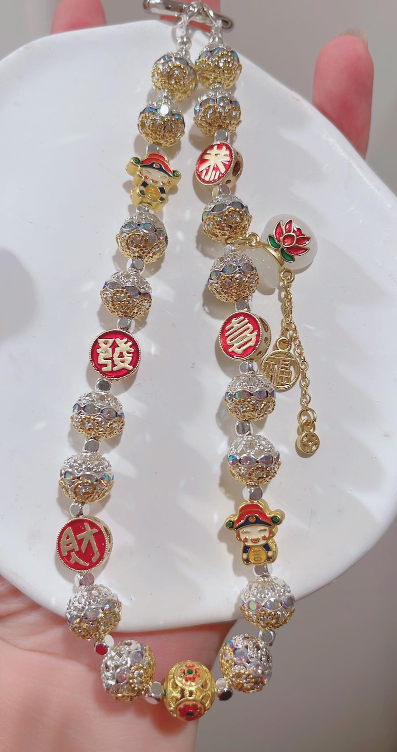 May include: A gold bracelet with decorative beads and charms. The bracelet features round, textured beads interspersed with smaller, crystal-studded spheres. Charms include a red and gold emblem, a cartoon figure, and a lotus flower.