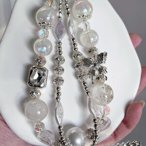 May include: A beaded keychain with multiple strands of white and iridescent beads. The strands include clear, round, and faceted beads, along with silver accents and a silver butterfly charm. The keychain is attached to a silver clasp.