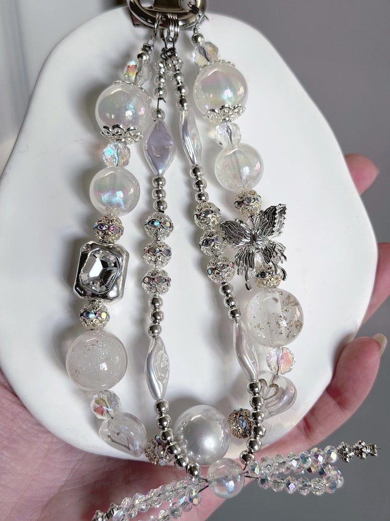 May include: A beaded keychain with multiple strands of white and iridescent beads. The strands include clear, round, and faceted beads, along with silver accents and a silver butterfly charm. The keychain is attached to a silver clasp.