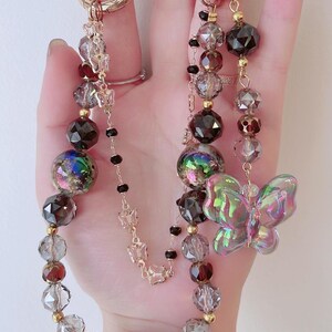 May include: Two beaded keychains with gold-colored rings. One keychain features a clear iridescent butterfly charm. Both keychains have a mix of faceted and round beads in shades of brown, clear, and iridescent. Small gold butterfly charms are also present.
