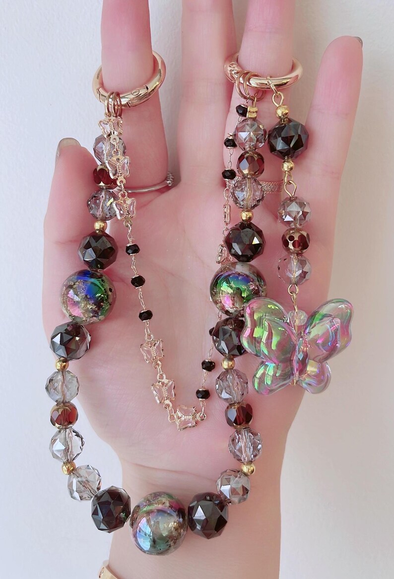 May include: Two beaded keychains with gold-colored rings. One keychain features a clear iridescent butterfly charm. Both keychains have a mix of faceted and round beads in shades of brown, clear, and iridescent. Small gold butterfly charms are also present.