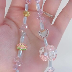 May include: A beaded keychain with pink and clear beads, gold accents, and a silver heart charm. The keychain features a lotus flower charm and a floral bead. The beads are strung on a silver chain.