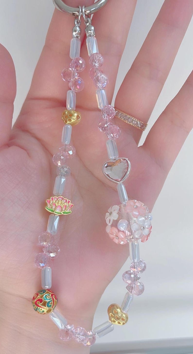 May include: A beaded keychain with pink and clear beads, gold accents, and a silver heart charm. The keychain features a lotus flower charm and a floral bead. The beads are strung on a silver chain.