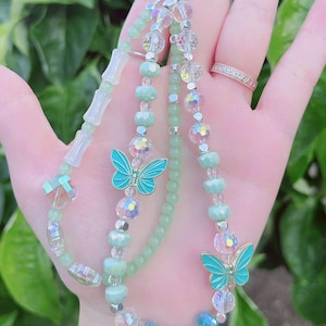 May include: A beaded keychain with a variety of beads in shades of green, clear, and iridescent. Two turquoise butterfly charms are featured. The keychain is held up by a silver-colored clasp. The background is blurred green foliage.