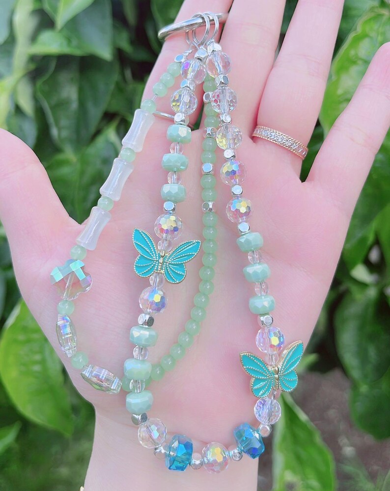 May include: A beaded keychain with a variety of beads in shades of green, clear, and iridescent. Two turquoise butterfly charms are featured. The keychain is held up by a silver-colored clasp. The background is blurred green foliage.