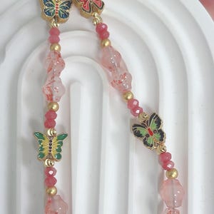 May include: A beaded keyring with a gold clasp and butterfly charms. The keyring features pink and gold beads, with colourful butterfly charms in red, blue, and green. A red fish charm is at the bottom.