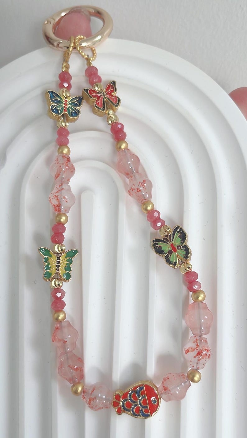 May include: A beaded keyring with a gold clasp and butterfly charms. The keyring features pink and gold beads, with colourful butterfly charms in red, blue, and green. A red fish charm is at the bottom.