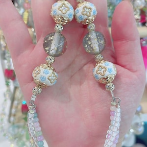 May include: A beaded necklace with a silver clasp. The necklace features white and gold beads with blue floral designs, clear beads, and gray beads with gold flecks. The lower portion of the necklace has a multi-colored beaded design.