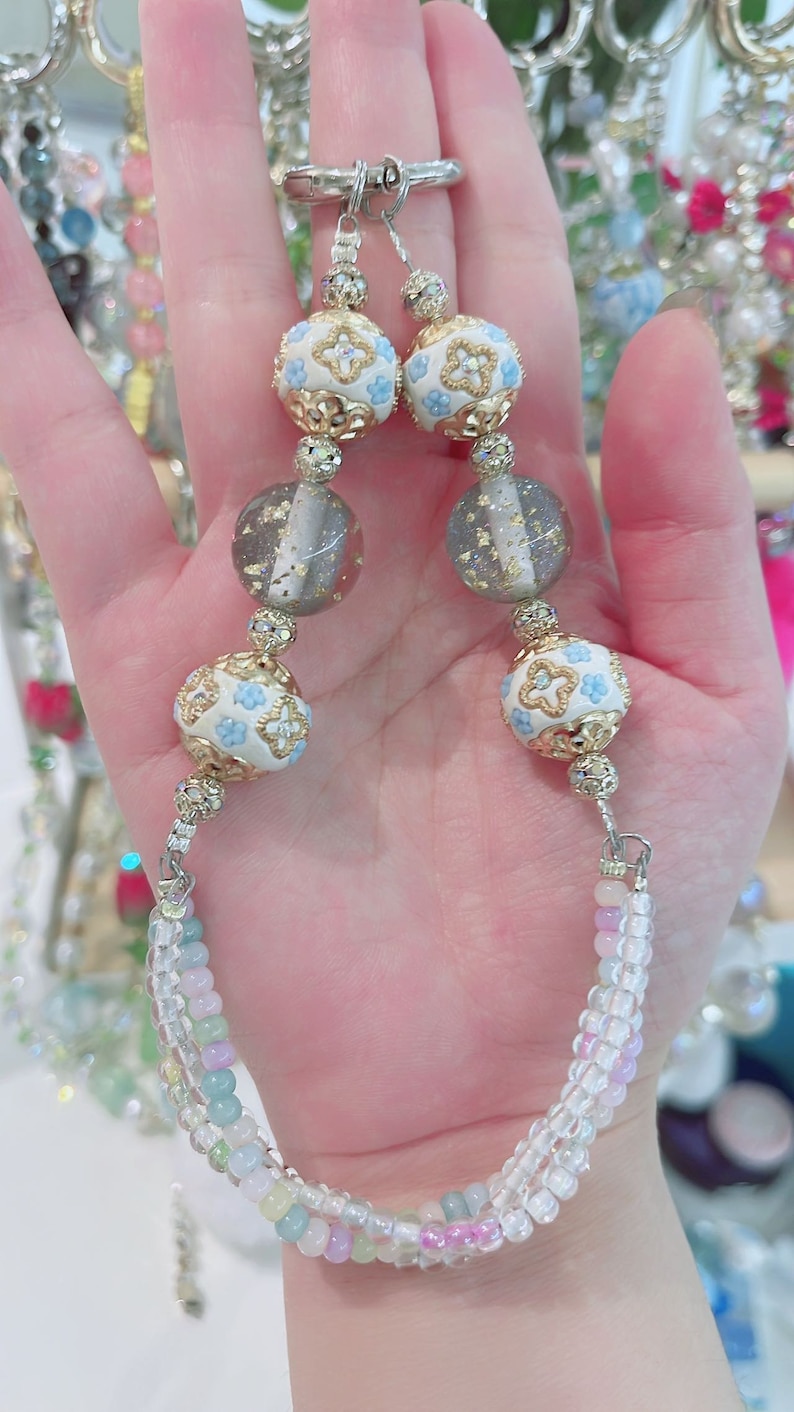 May include: A beaded necklace with a silver clasp. The necklace features white and gold beads with blue floral designs, clear beads, and gray beads with gold flecks. The lower portion of the necklace has a multi-colored beaded design.