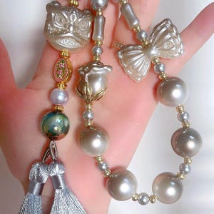 May include: A decorative keychain with a lion head, bow, and rose-shaped charm, embellished with pearls and gold details. The keychain includes silver tassels and various pearl sizes.