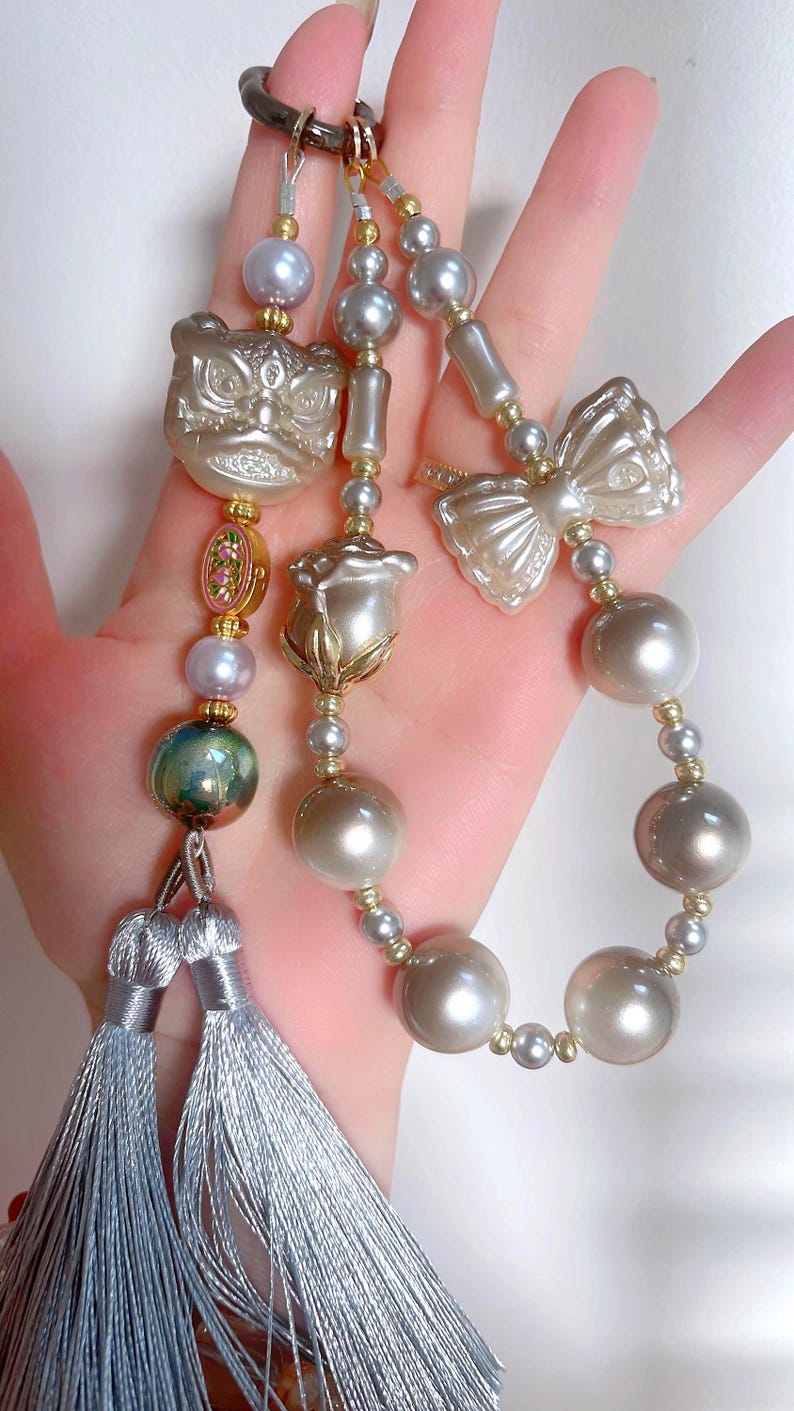 May include: A decorative keychain with a lion head, bow, and rose-shaped charm, embellished with pearls and gold details. The keychain includes silver tassels and various pearl sizes.
