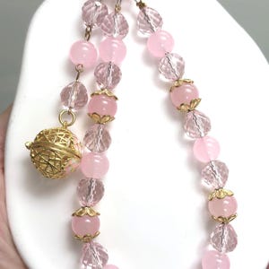 May include: A pink beaded necklace with a gold charm and a pink clip. The necklace features alternating pink and clear crystal beads. The charm is a gold filigree sphere. The clip is attached to a pink ring.