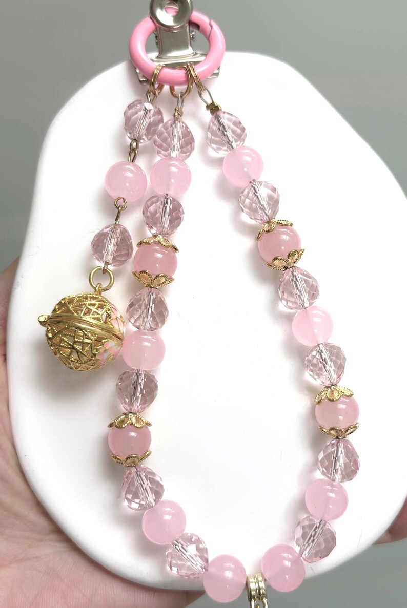 May include: A pink beaded necklace with a gold charm and a pink clip. The necklace features alternating pink and clear crystal beads. The charm is a gold filigree sphere. The clip is attached to a pink ring.