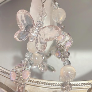 May include: A delicate, dangling earring featuring a butterfly and orb design. The earring is made of clear, iridescent, and pink-toned beads and crystals, suspended from a silver-colored hoop.