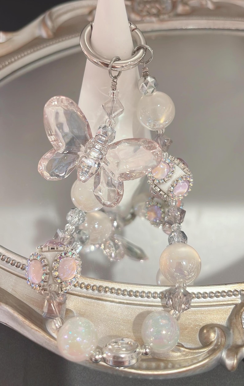 May include: A delicate, dangling earring featuring a butterfly and orb design. The earring is made of clear, iridescent, and pink-toned beads and crystals, suspended from a silver-colored hoop.