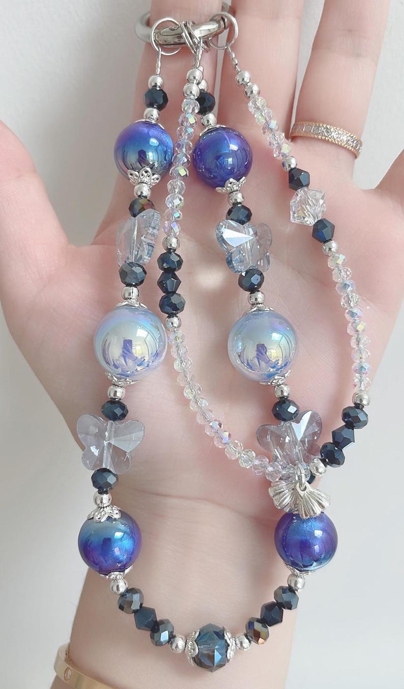 May include: A beaded keychain featuring iridescent blue and silver beads, clear faceted beads, and butterfly charms. The keychain has a silver clasp and is designed with multiple strands of beads.