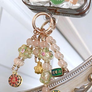 May include: A gold-coloured keychain with a beaded charm. The charm features pink beads, floral accents, and decorative pendants. The keychain is attached to a phone case with Chinese characters and a "Happy" sticker.