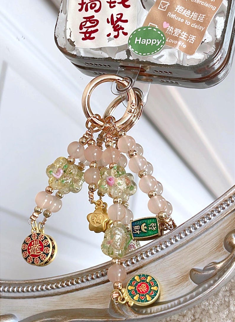 May include: A gold-coloured keychain with a beaded charm. The charm features pink beads, floral accents, and decorative pendants. The keychain is attached to a phone case with Chinese characters and a "Happy" sticker.