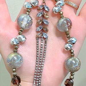 May include: A necklace featuring a silver-tone chain and an assortment of beads. The beads include faceted crystals, iridescent orbs, and dark brown, faceted beads. A central pendant showcases a leaf design with clear and amber crystals and dangling rhinestone strands.