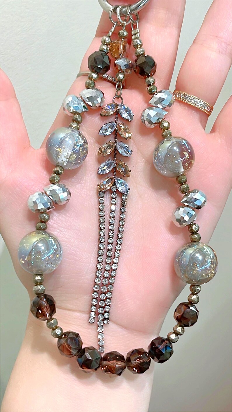 May include: A necklace featuring a silver-tone chain and an assortment of beads. The beads include faceted crystals, iridescent orbs, and dark brown, faceted beads. A central pendant showcases a leaf design with clear and amber crystals and dangling rhinestone strands.