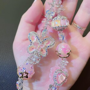 May include: A beaded keychain with a silver clasp. The keychain features a butterfly, heart, and star-shaped charms, along with pink and clear beads. The charms are embellished with rhinestones and crystals, creating a sparkling effect.
