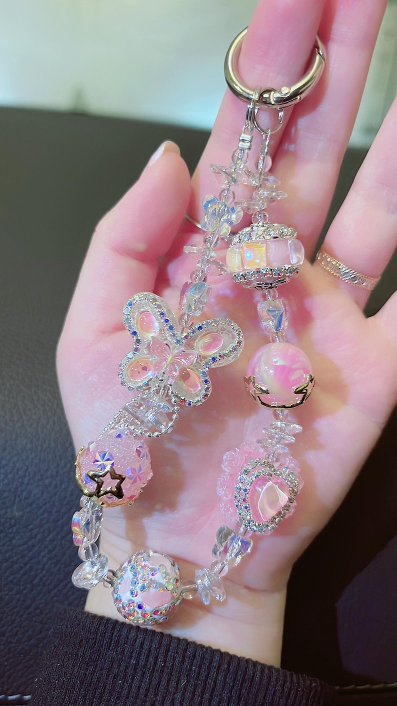 May include: A beaded keychain with a silver clasp. The keychain features a butterfly, heart, and star-shaped charms, along with pink and clear beads. The charms are embellished with rhinestones and crystals, creating a sparkling effect.