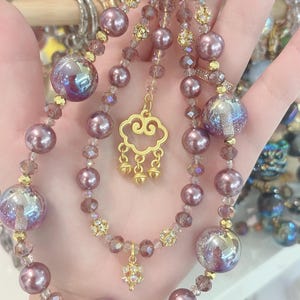 May include: A decorative beaded keychain with a gold clasp. The keychain features purple beads, iridescent orbs, and gold accents. A gold cloud charm and a small floral charm dangle from the beaded strands.