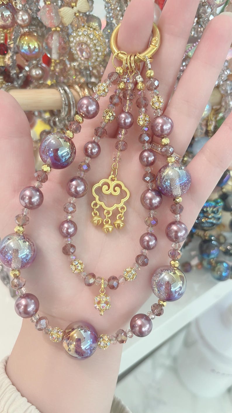 May include: A decorative beaded keychain with a gold clasp. The keychain features purple beads, iridescent orbs, and gold accents. A gold cloud charm and a small floral charm dangle from the beaded strands.