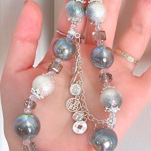 May include: A beaded necklace with iridescent grey and silver spheres, clear and grey crystal beads, and small silver charms. The necklace is held in a hand, highlighting its intricate design and craftsmanship.