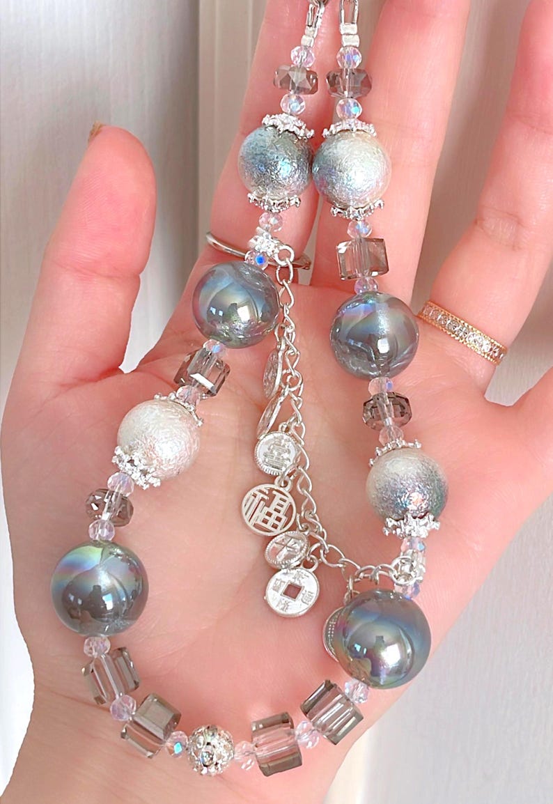 May include: A beaded necklace with iridescent grey and silver spheres, clear and grey crystal beads, and small silver charms. The necklace is held in a hand, highlighting its intricate design and craftsmanship.