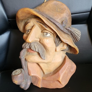 May include: A wooden wall sculpture of a man with a mustache and a hat, holding a pipe in his mouth. The sculpture is painted in shades of brown, beige, and black.