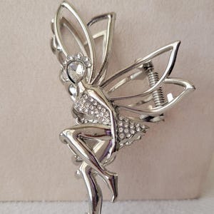 May include: Silver fairy hair clip with crystal embellishments. The fairy is sitting with her wings spread out.