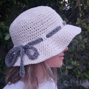 Puede incluir: Un sombrero de sol de ganchillo color crema con ala ancha. El sombrero presenta una banda gris y un lazo decorativo. Un accesorio elegante para la protección solar y añade un toque de encanto artesanal.