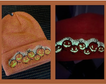 Puede incluir: Gorro de punto naranja con una fila de caras de calabaza talladas bordadas. El gorro brilla en la oscuridad, con las caras de calabaza talladas brillando en verde.
