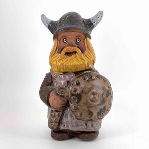 May include: A ceramic figurine of a Viking warrior, featuring a horned helmet, a yellow beard, and a shield. The figure is painted in shades of brown, gray, and yellow, with a detailed design. The figurine is standing upright.