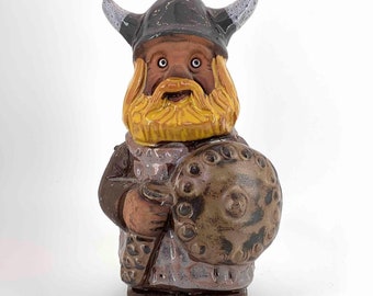 May include: A ceramic figurine of a Viking warrior, featuring a horned helmet, a yellow beard, and a shield. The figure is painted in shades of brown, gray, and yellow, with a detailed design. The figurine is standing upright.
