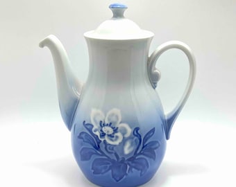 Puede incluir: Una cafetera de porcelana blanca con un diseño floral azul y blanco. La cafetera tiene un cuerpo redondeado y una tapa con un pomo. El diseño floral presenta una flor blanca con hojas azules.