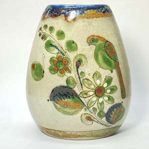 May include: Cream-colored ceramic vase with a blue rim and floral and bird design. The vase features a green bird, green flowers, and a butterfly. The design includes green, orange, and brown accents. The vase has a rounded shape.