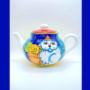 May include: A white ceramic teapot with a blue, yellow, and orange cat design. The teapot has a red lid with white polka dots and a white spout. The cat is holding a yellow flower pot with orange flowers.