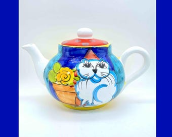 Puede incluir: Una tetera de cerámica blanca con un diseño de gato azul, amarillo y naranja. La tetera tiene una tapa roja con lunares blancos y un pico blanco. El gato sostiene una maceta amarilla con flores naranjas.