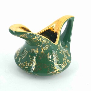 Puede incluir: Una jarra de crema de cer&aacute;mica verde con un patr&oacute;n floral dorado y un asa dorada. La jarra tiene forma de p&aacute;jaro con un pico ancho.